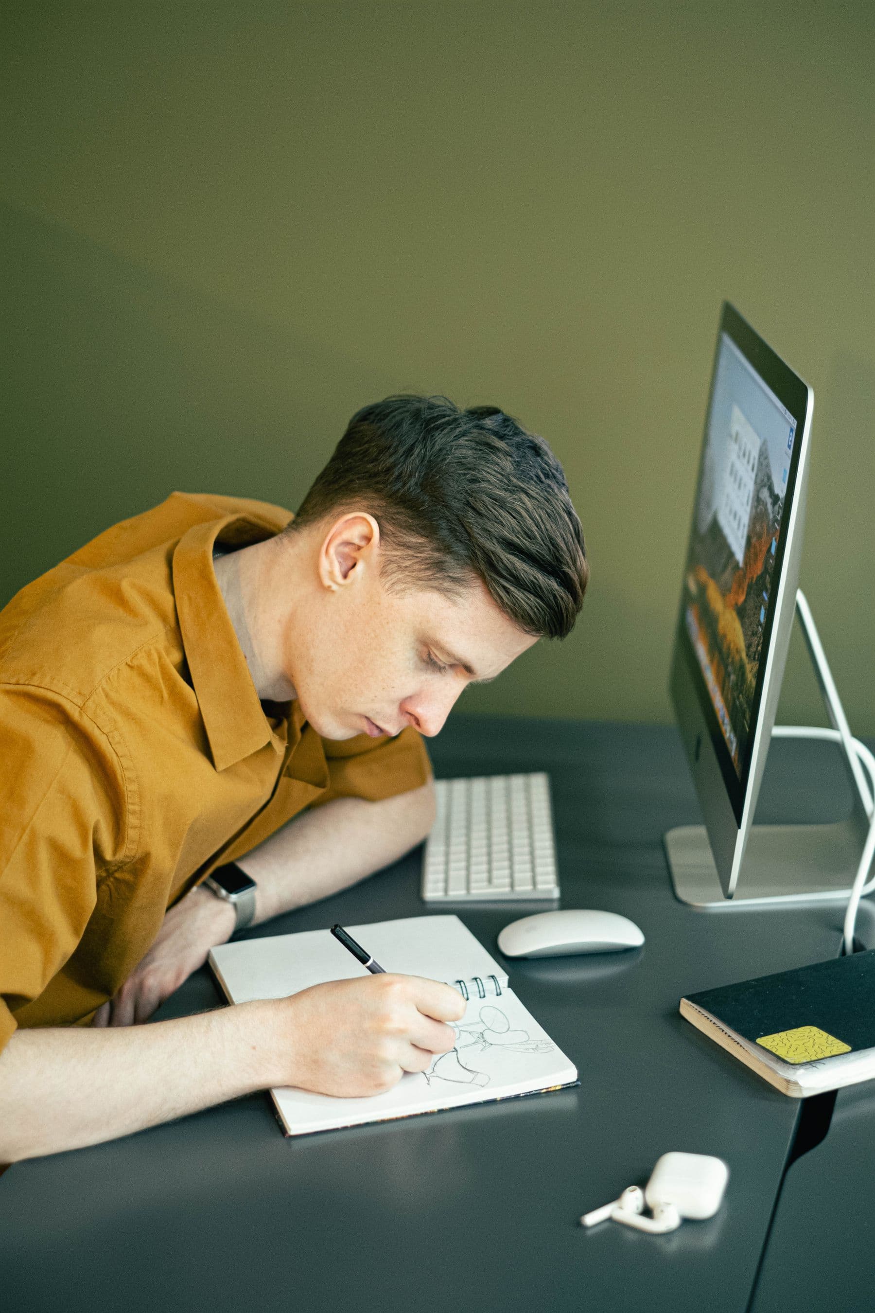 A man sketching in a notebook beside his desktop computer.