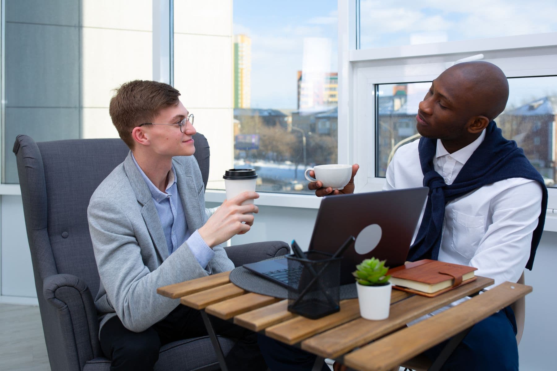 Two men talking over coffee beside a laptop in a bright office.