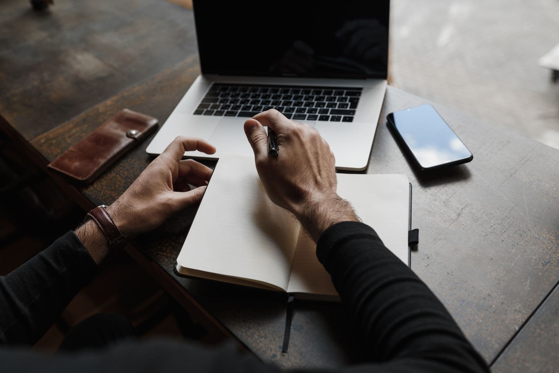 Hands taking notes in a notebook beside a laptop and phone.